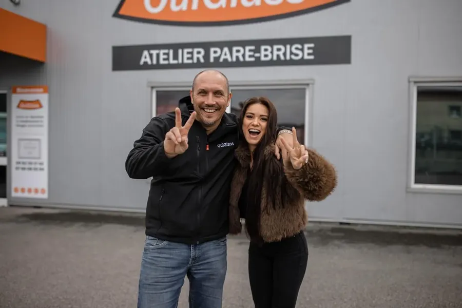 Deux personnes sourient et font le signe de la victoire devant l'enseigne « Atelier pare-brise » d'un centre OuiGlass
